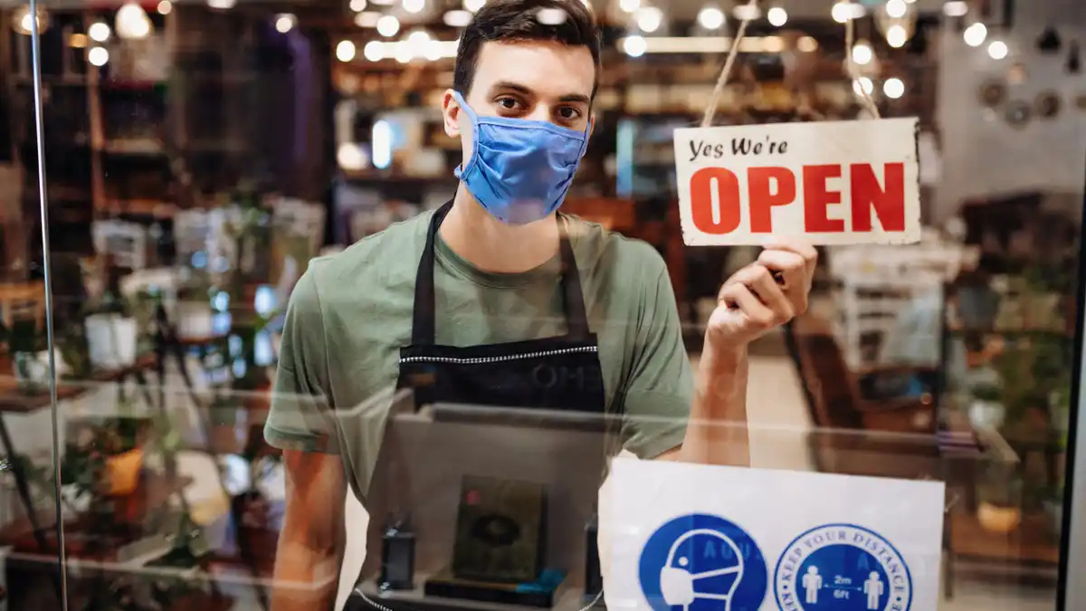 server_wearing_face_mask_and_holding_Open_sign_in_cafe_faby5i.jpg